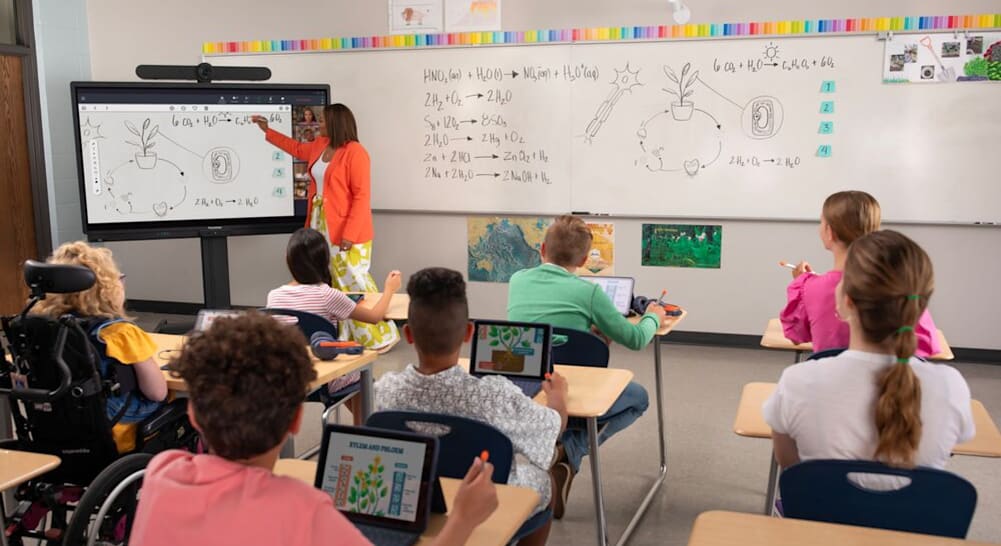 Students in a classroom using tablets while a teacher explains a plant diagram on a digital whiteboard.