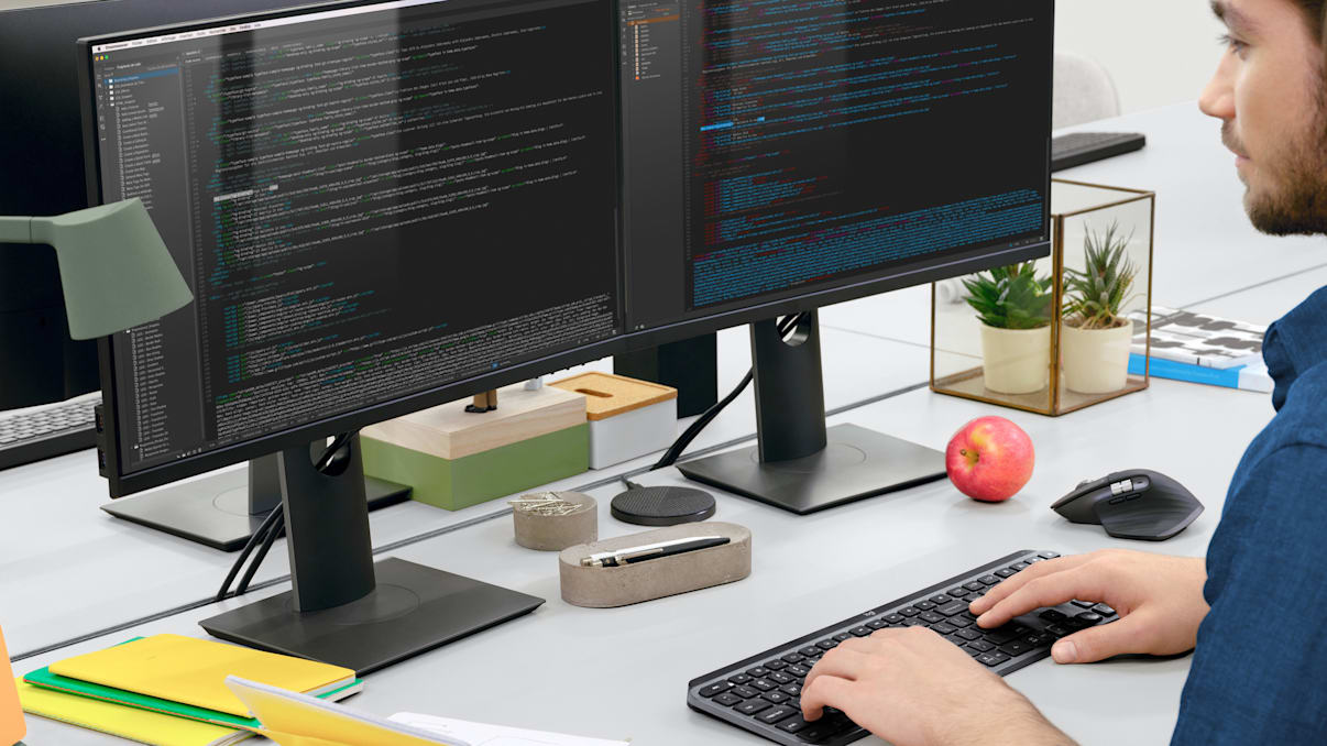 A programmer codes on dual monitors using his MX Keys for Business keyboard.