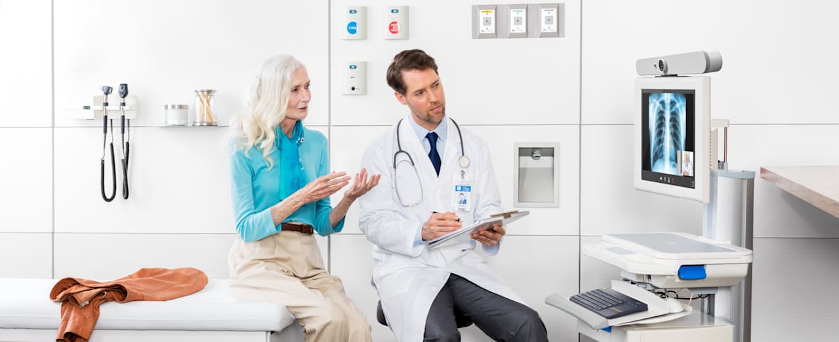 Doctor consulting patient with Logitech healthcare video system and digital X-ray on medical cart monitor.