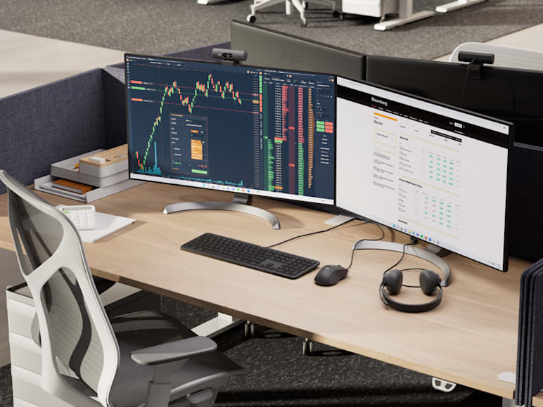 Modern shared desk setup with dual ultra-wide monitors displaying financial data, in an open office environment.