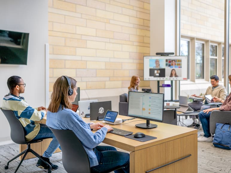 In an open office, some people work at desks using computers with Logitech headsets and accessories, while others sit in the background attending a video conference on a large screen.