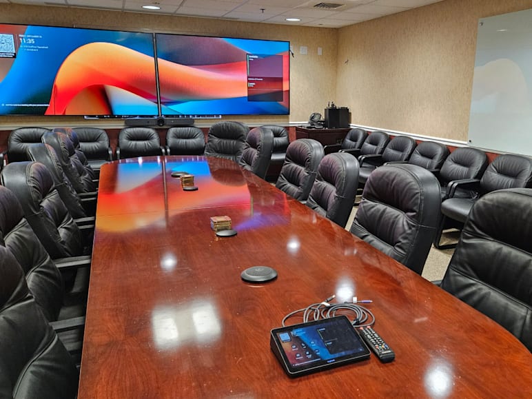 Conference room with long wooden table, leather chairs, dual display screens, and Logitech video conferencing setup