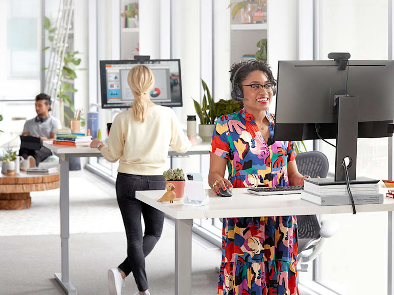 In a bright, modern office, two women work at standing desks using various Logitech accessories, while others relax in the background.