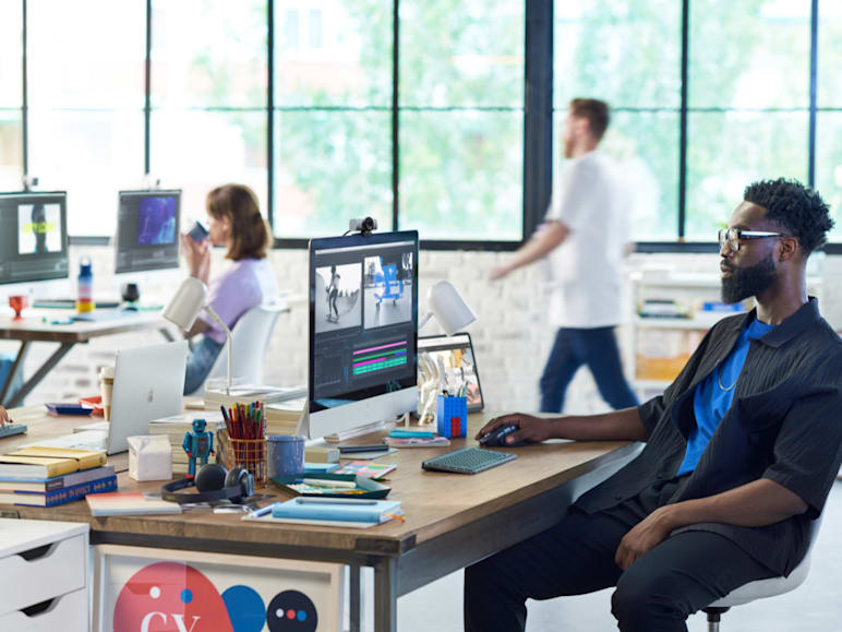 Modern open office with shared desks, where a man works on a desktop computer. The workspace includes a Logitech webcam, keyboard, mouse, headset and creative tools