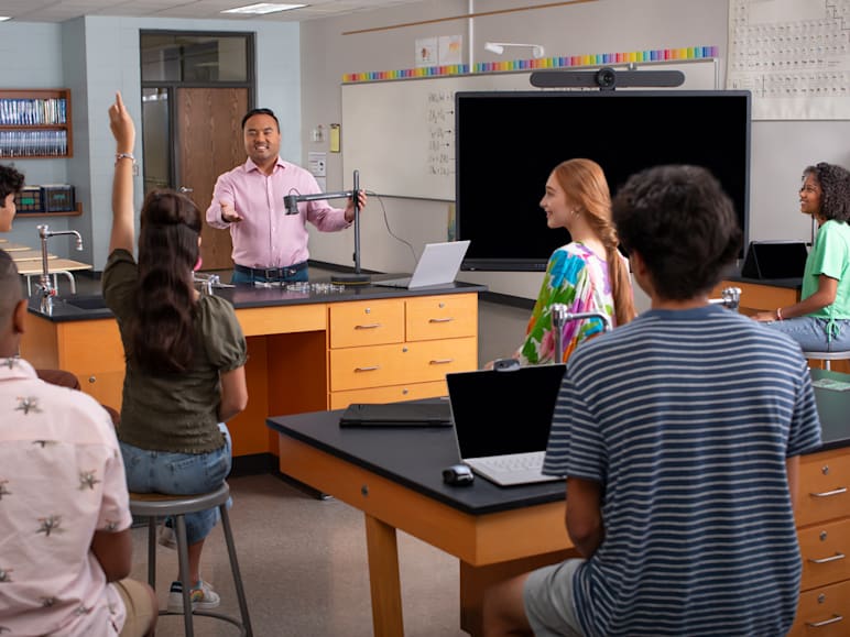 Teacher in a classroom using Logitech video conferencing system, engaging students at lab desks with laptops and science equipment.