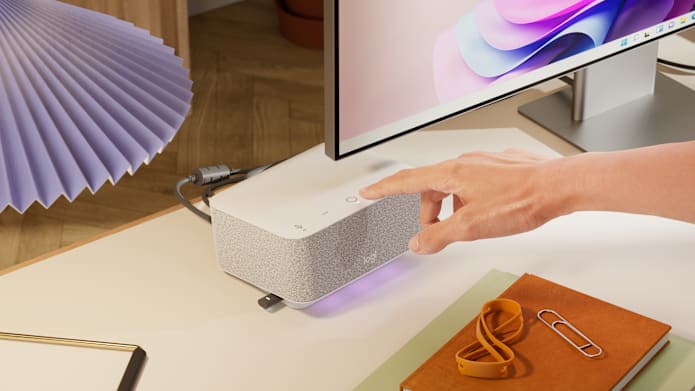Hand pressing button on Logitech Dock placed on office desk next to monitor, notebook, and accessories.