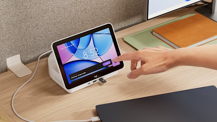 Hand touching Logitech Dock Flex display on office desk to join a meeting, showing time, room name, and meeting details on screen.